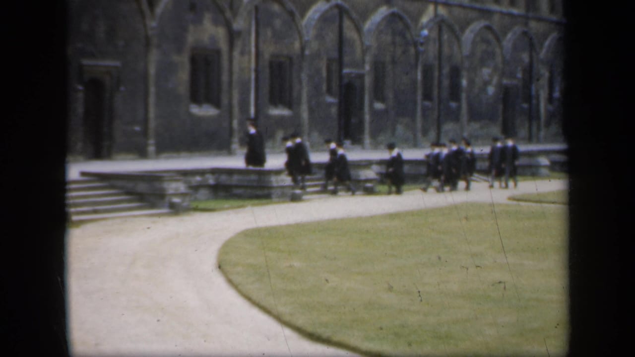 Robed figures walk across a stone courtyard beside steps and a lawn.