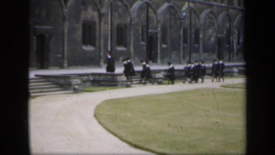In England 1949 home-movie footage, people in black academic robes cross a courtyard near stone steps and arched windows.