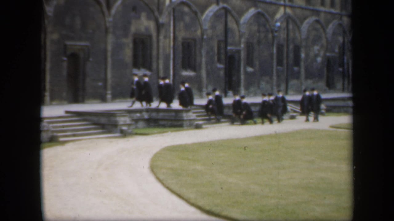 Robed academic figures gather and walk near stone steps in front of a historic-looking English university building.