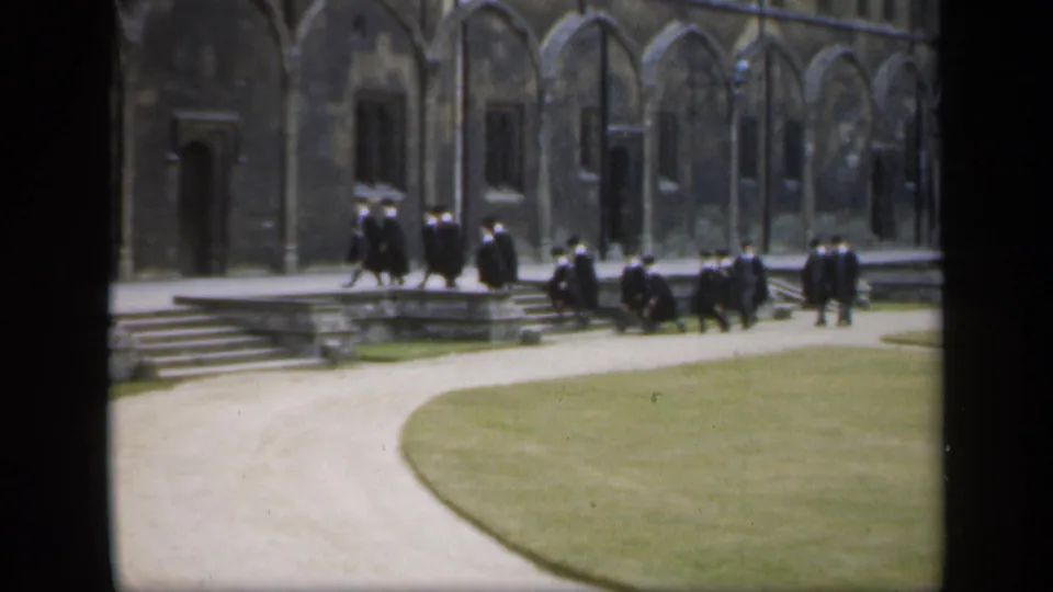 Robed academic figures gather and walk near stone steps in front of a historic-looking English university building.