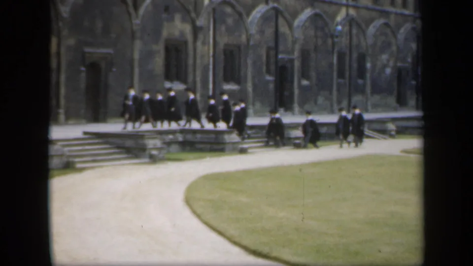 People in black academic robes move across broad stone steps with a Gothic-style facade and lawn visible in the frame.