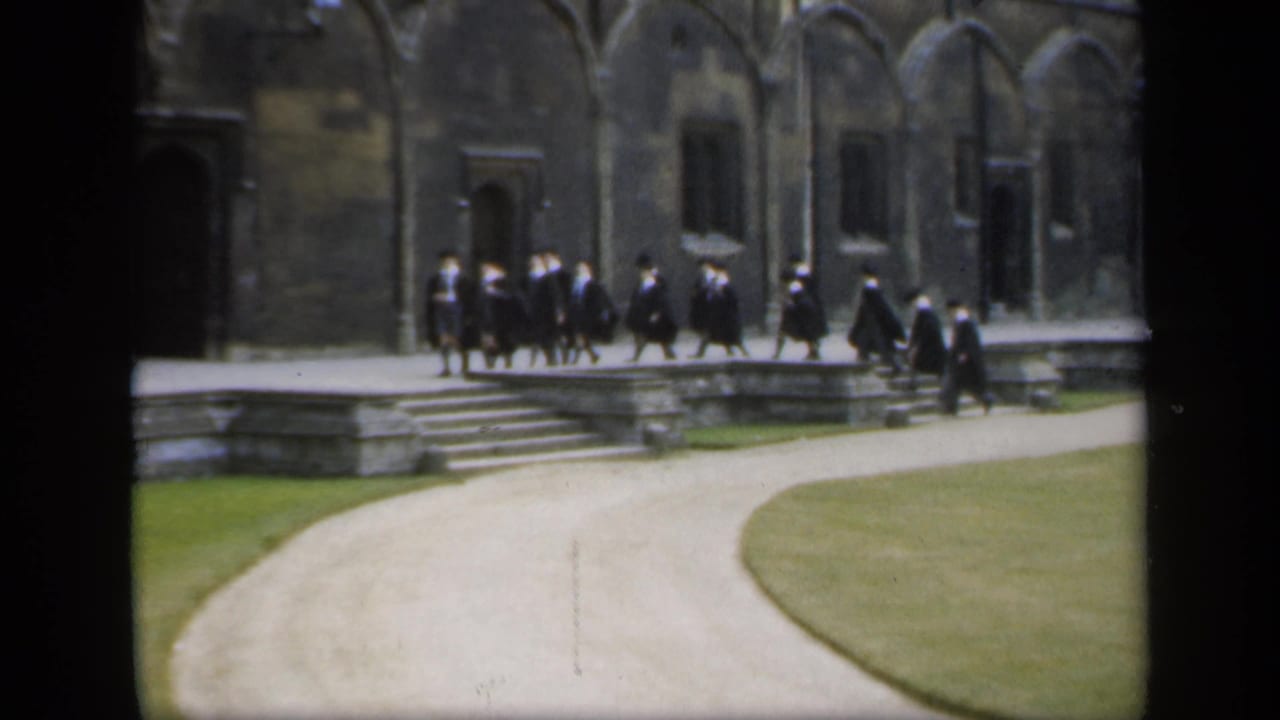 Robed men walk across a raised stone walkway beside a historic courtyard building.