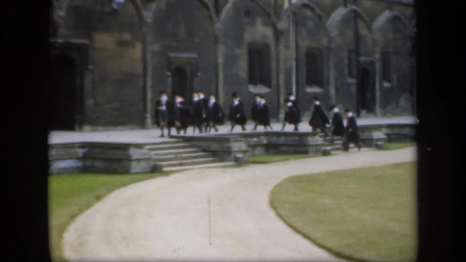 In 1949 England, men in academic robes move along a raised stone walk beside Gothic-style arches and a curved path.