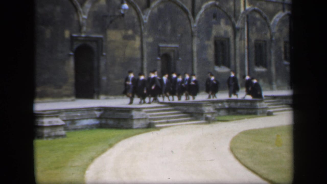 In 1949 England, a robed group walks in formation past stone steps, arched masonry, and dark doorways on a campus courtyard.
