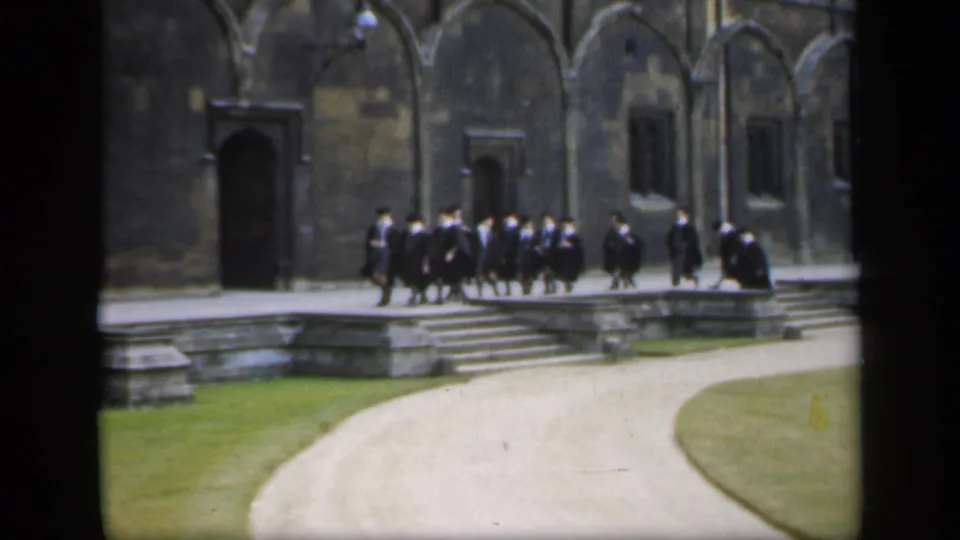 In 1949 England, a robed group walks in formation past stone steps, arched masonry, and dark doorways on a campus courtyard.