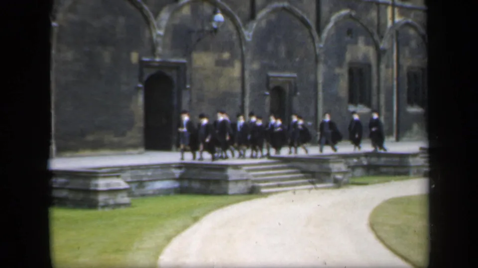 In 1949 England, men in academic dress continue across the raised stone edge of a courtyard near a large doorway and lawn.