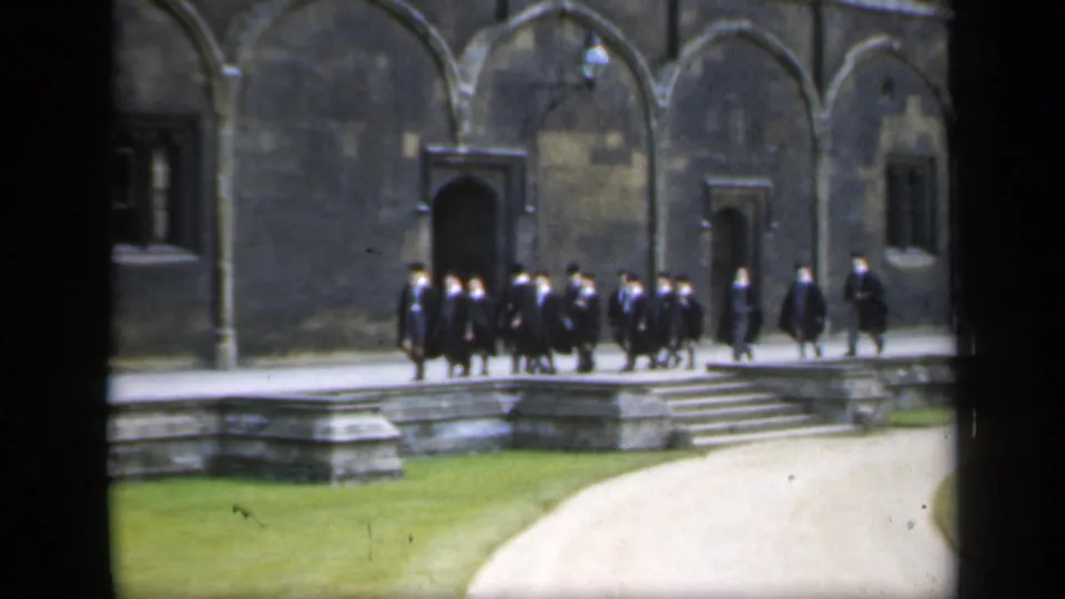 In 1949 England, the academic-robed group crosses a historic stone courtyard with trailing figures near windows and steps.