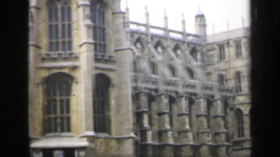 1949 England frame showing a side section of an ornate Gothic stone facade with tall windows, buttress details, and pale overcast light.