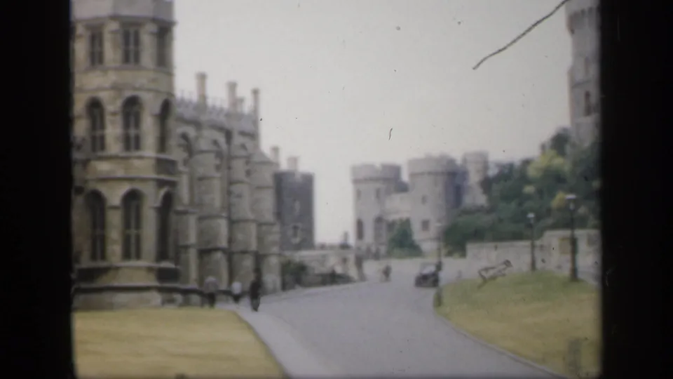1949 England frame with Gothic stone architecture at left, a curving road, lawn, tree, and small distant visitors.