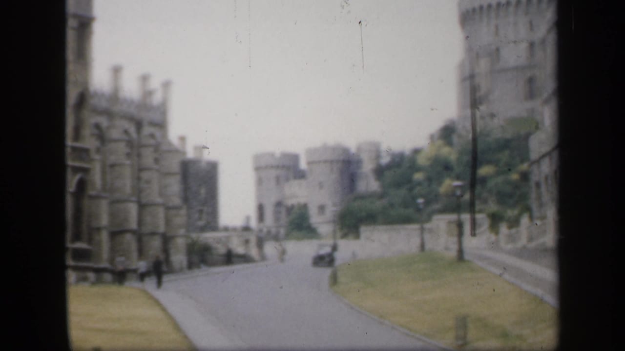Stone castle towers above a quiet curving road with grass verges and distant figures.