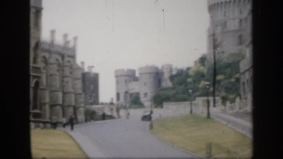 1949 England home-movie frame showing a stone castle complex beside a quiet road, with street lamps, grass, and distant pedestrians.
