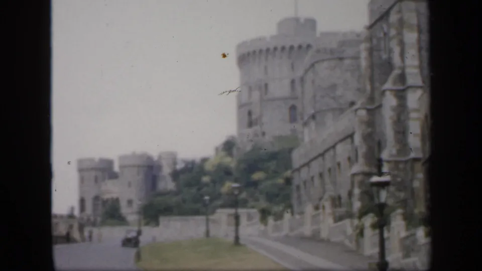 Soft 8mm frame of castle masonry and a roadside approach in England, with a tall tower, wall, lamps, and faint distant figures.