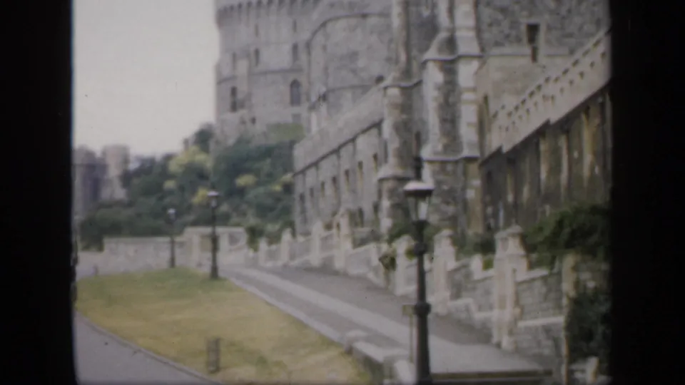 Final 1949 England frame showing the castle wall and tower beside a quiet road, with lamps, grass, and soft film grain.