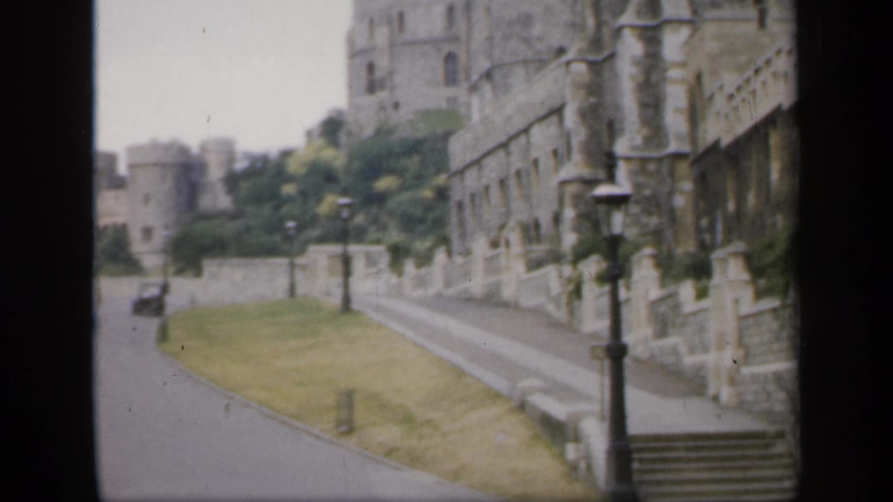 1949 England home-movie view of a road running below historic stone castle walls with grass, lampposts, and trees.