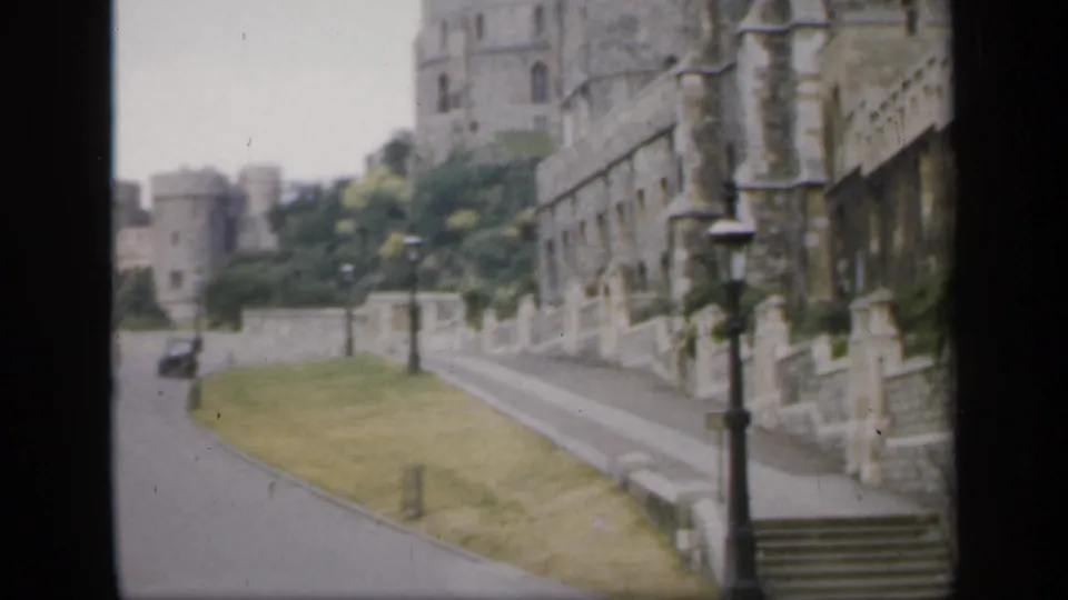 1949 England home-movie view of a road running below historic stone castle walls with grass, lampposts, and trees.