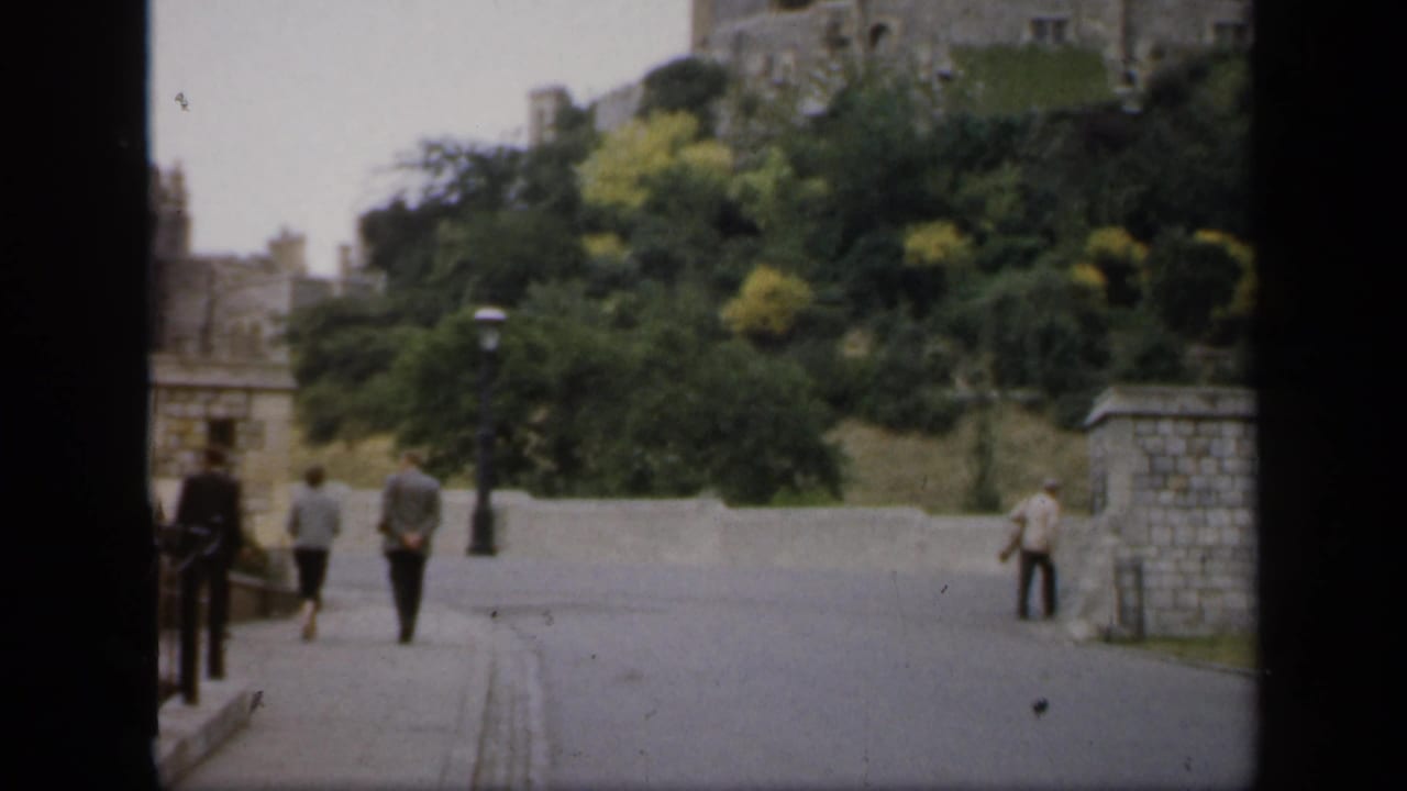 1949 England castle approach with several pedestrians walking near stone walls, lampposts, trees, and a road.