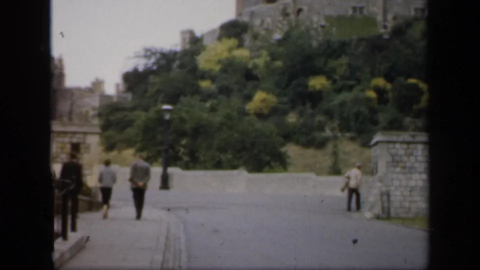 1949 England castle approach with several pedestrians walking near stone walls, lampposts, trees, and a road.