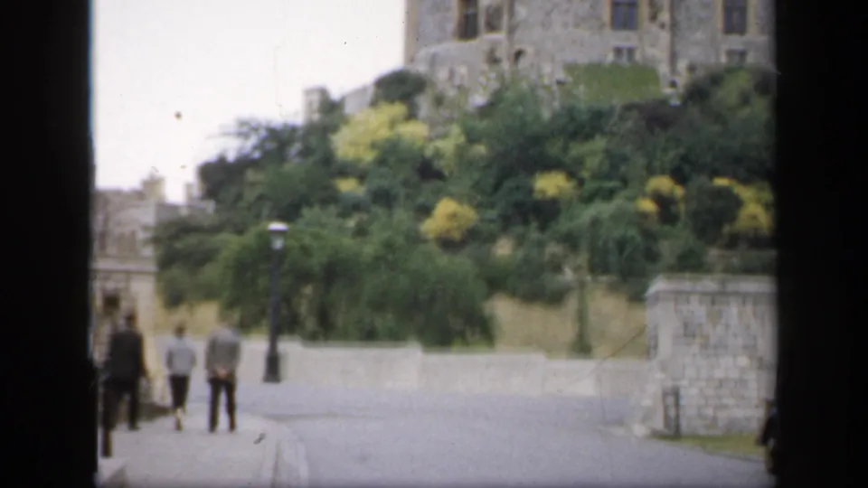 1949 England home-movie frame showing distant walkers on a road below castle stonework and thick greenery.