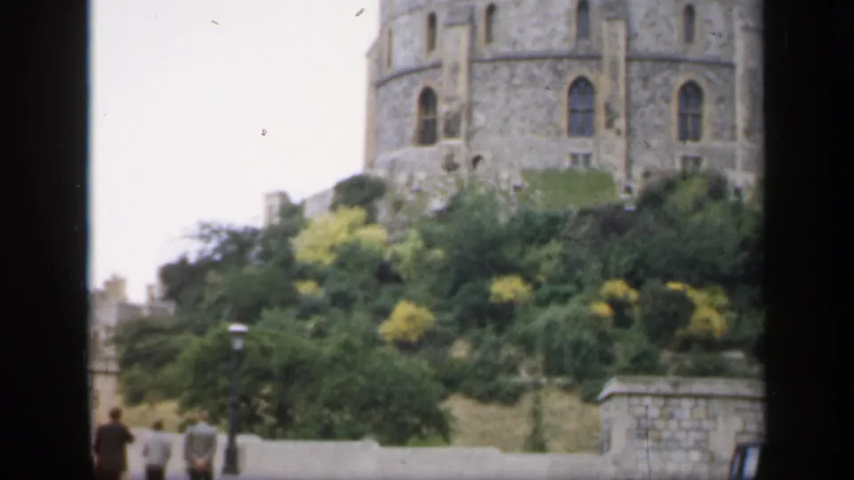 1949 England castle view with a tower rising over trees, a road below, and distant pedestrians near the wall.