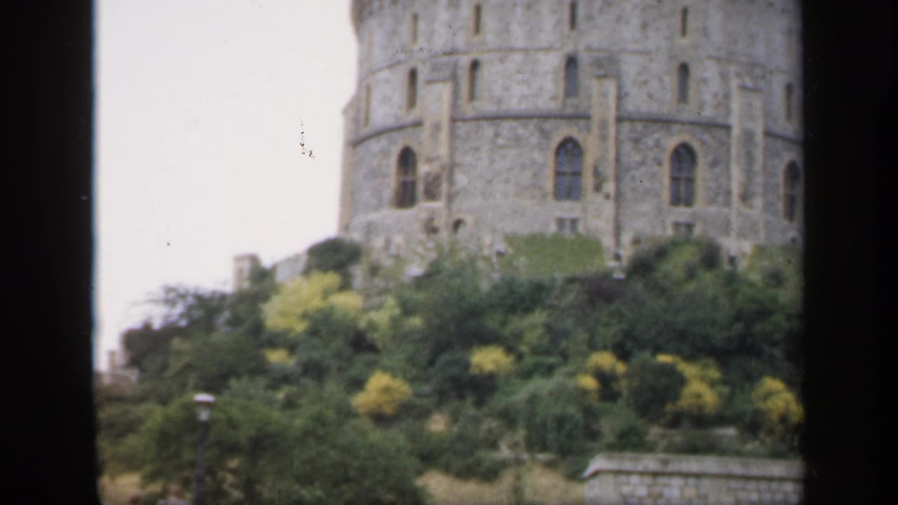 1949 England home-movie frame showing a round stone castle tower rising above garden shrubs, partially framed by a dark opening.
