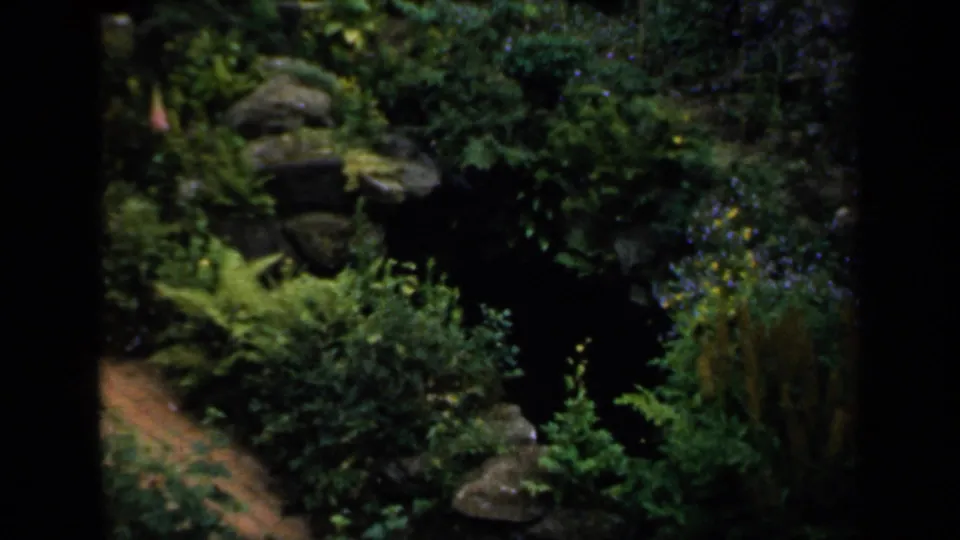 1949 England garden view inside castle grounds, with rockwork, shrubs, and a path seen through dark foreground framing.