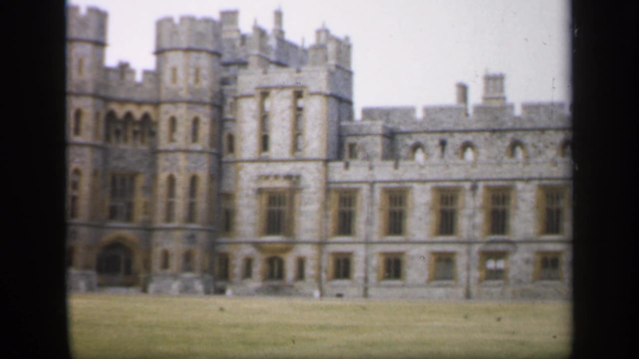 1949 England exterior frame showing the stone castle frontage, crenelated roofline, paired towers, arched opening, windows, and open lawn.