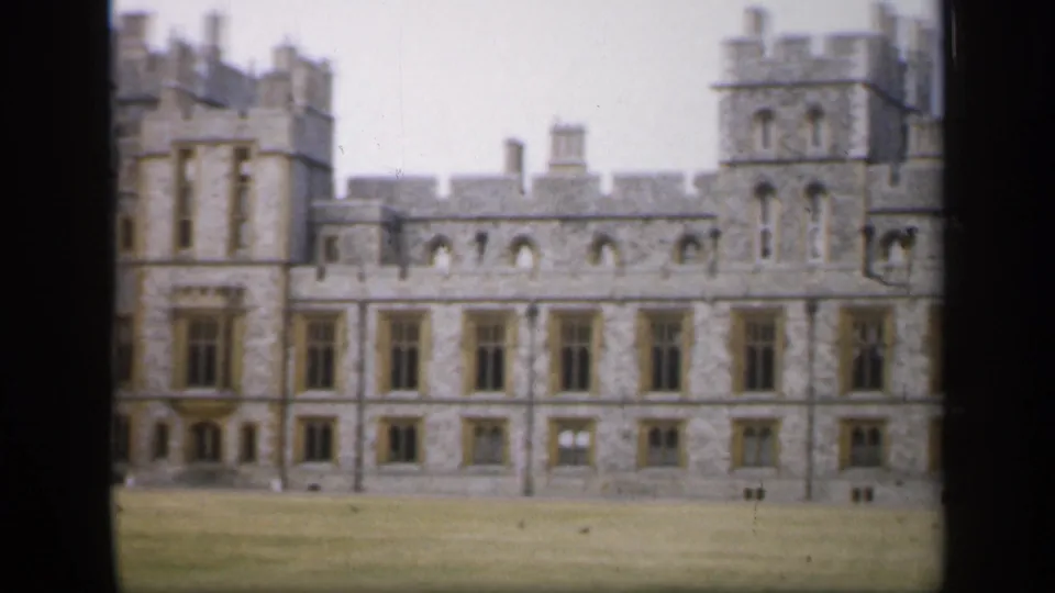1949 England 8mm frame of the castle's long stone wall, crenelated roofline, brown-framed windows, part of a tower, and grassy foreground.