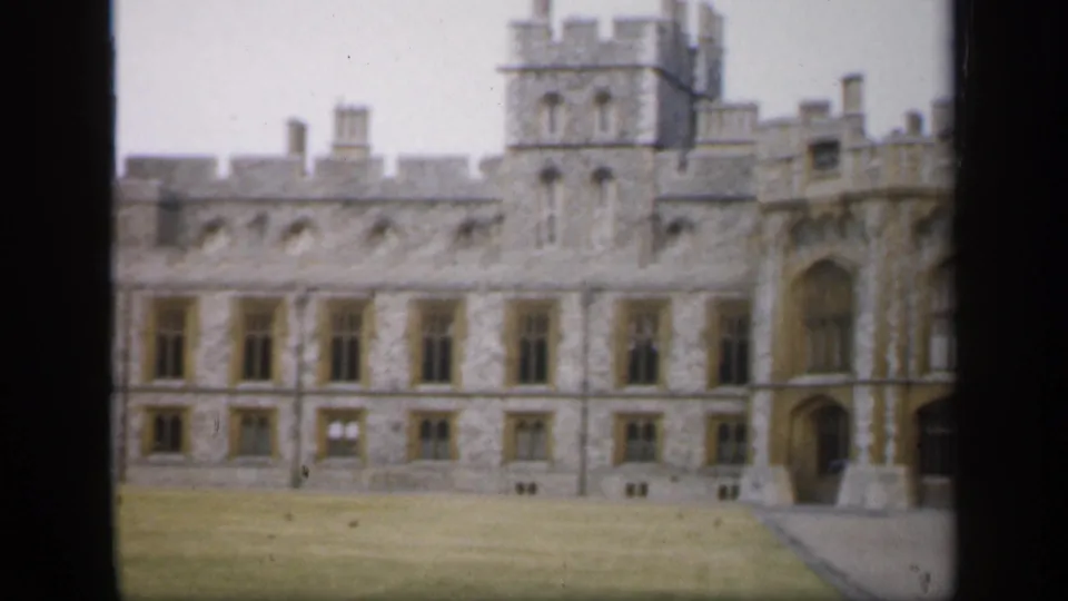 1949 England exterior frame showing a castle wing with repeated windows, a tower, battlements, arched passage, lawn, and walkway.