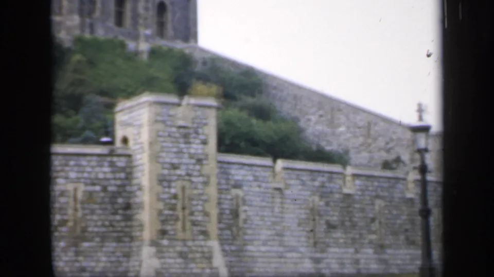 1949 England frame showing the castle perimeter wall, sloped stonework, green planting, cloudy sky, and a lamp post at right.