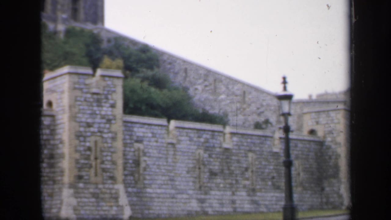 1949 England 8mm frame showing a stone fortification wall, black lamppost, green lawn, and pale overcast sky.