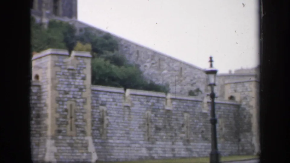 1949 England 8mm frame showing a stone fortification wall, black lamppost, green lawn, and pale overcast sky.