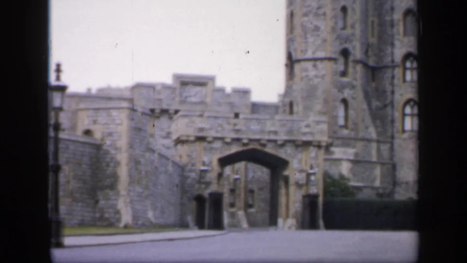 1949 England 8mm frame showing a castle-like arched entrance, adjoining tower, paved road, lamppost, and dark hedge.