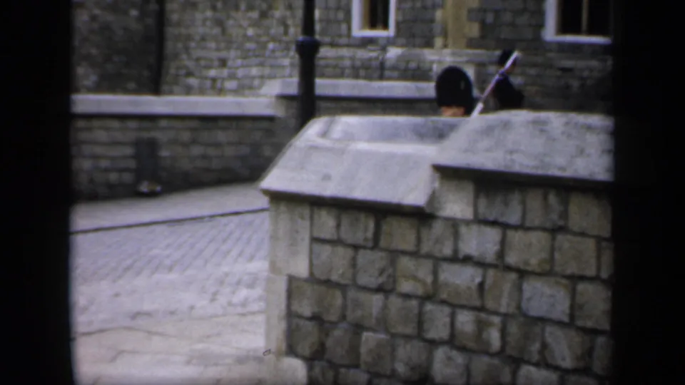 1949 England home-movie frame showing a guard's black hat and rifle partly visible above a stone wall in a courtyard.