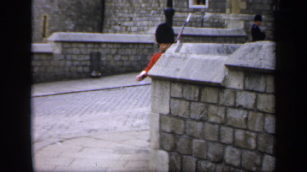 Guard in a red tunic walking beside a stone wall with a rifle.