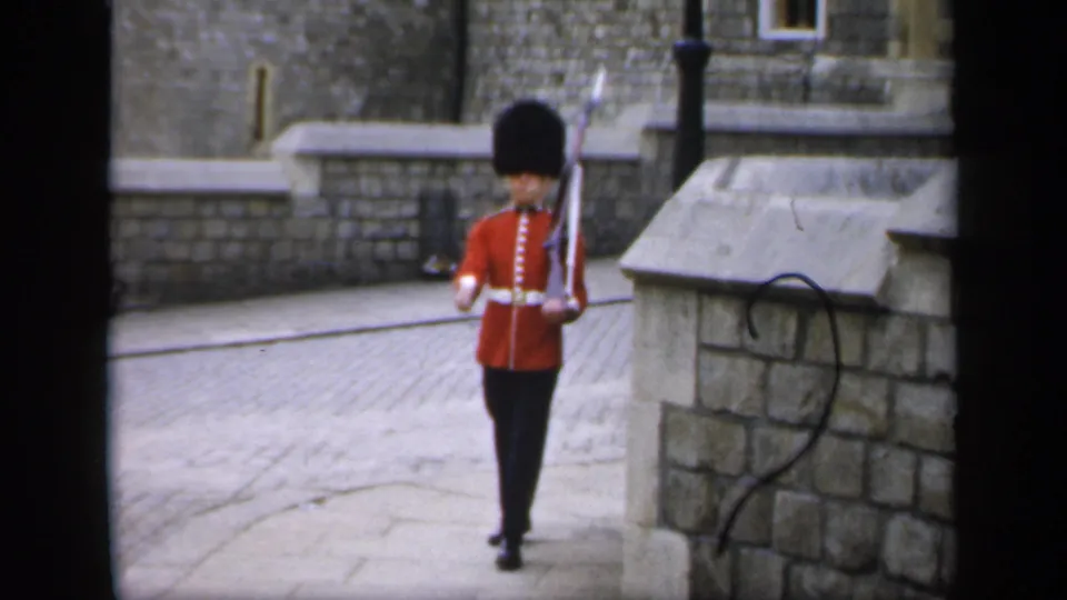 1949 England home-movie frame showing a red-coated guard from behind, carrying a rifle near stone walls.