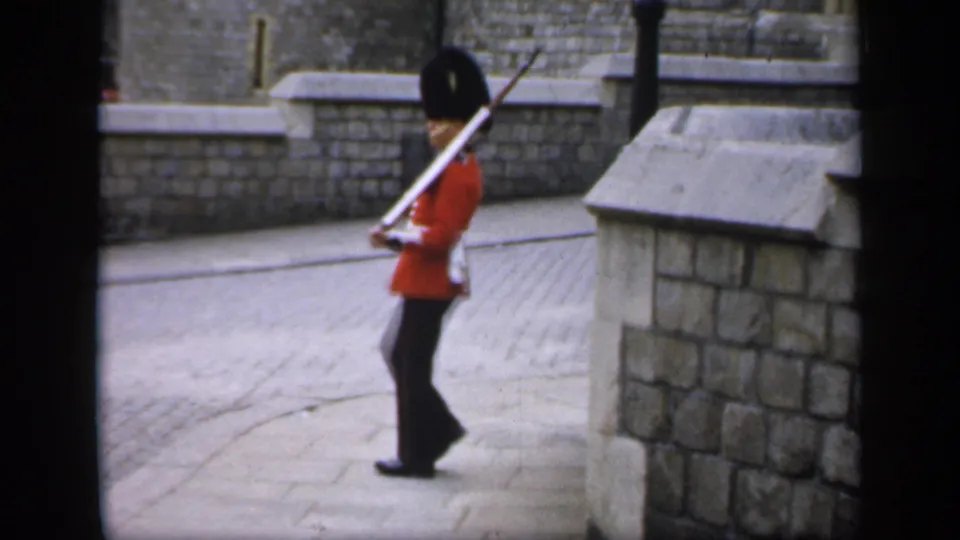 1949 England frame with a side view of a ceremonial guard in red tunic and tall black hat beside stone walls.