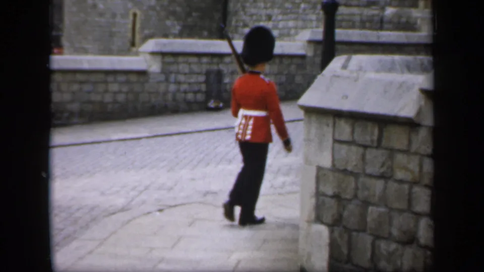 The red-coated guard turns near a low stone wall, with paved ground and historic masonry visible around him.