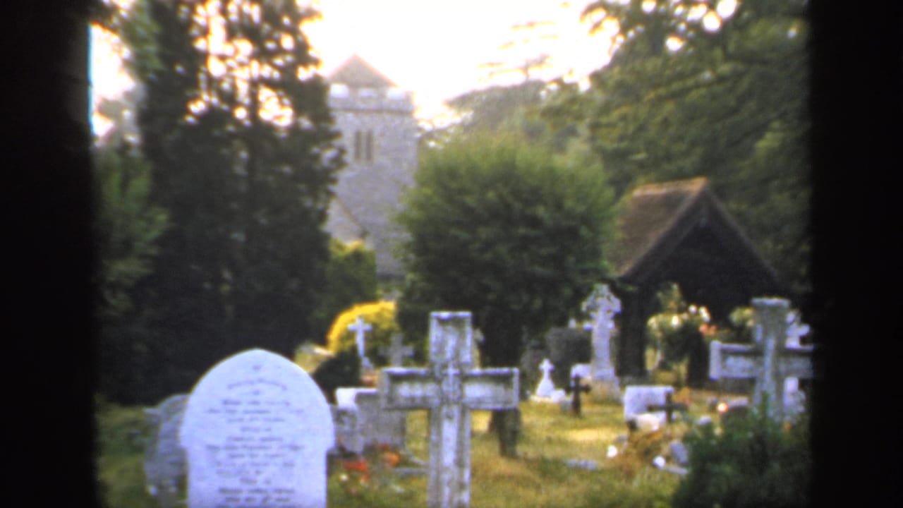 Churchyard seen through dark framing with white gravestones, trees, and a stone tower behind.