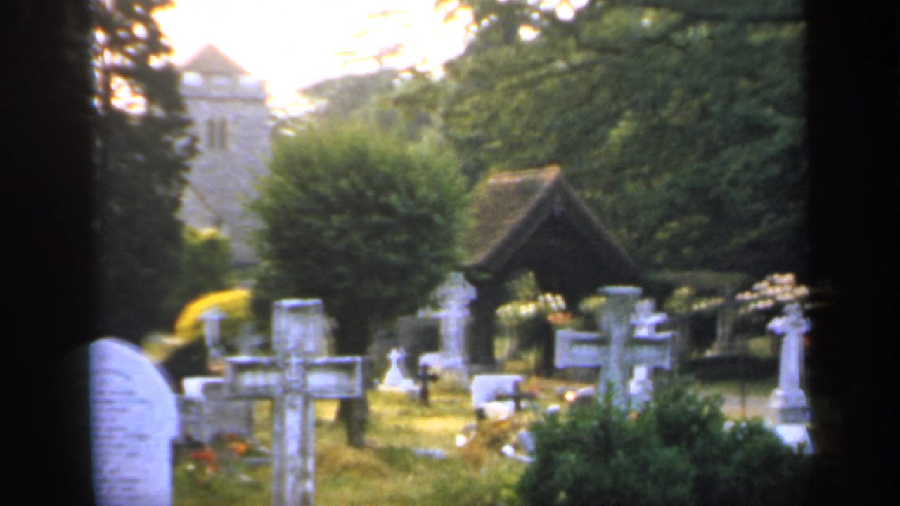 1949 England churchyard with rows of white gravestones, crosses, dense trees, a wooden shelter, and a stone church tower partly visible.
