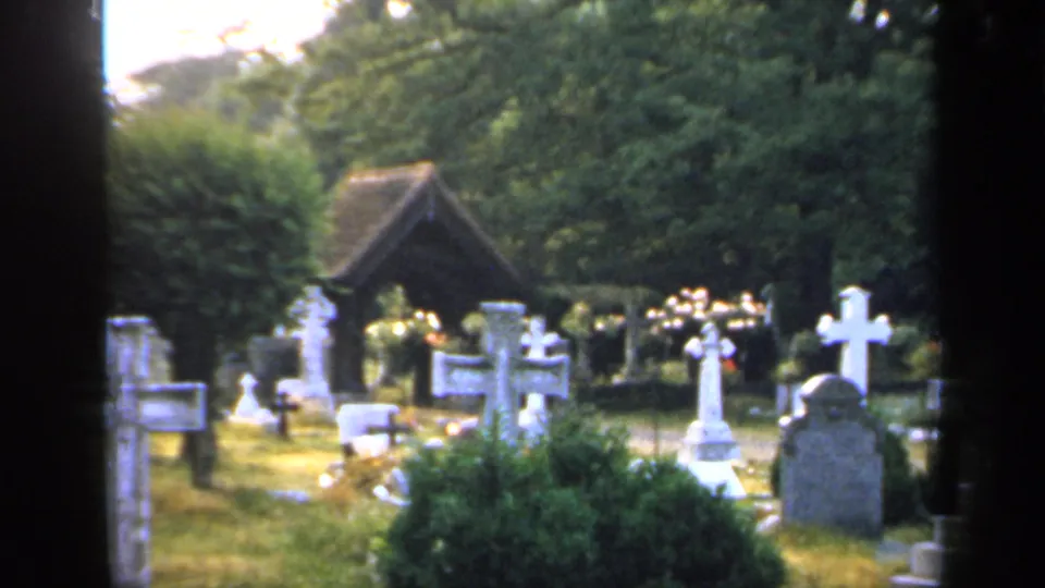 1949 England churchyard frame showing white crosses, headstones, shrubs, grass, and a small wooden shelter beneath dense trees.