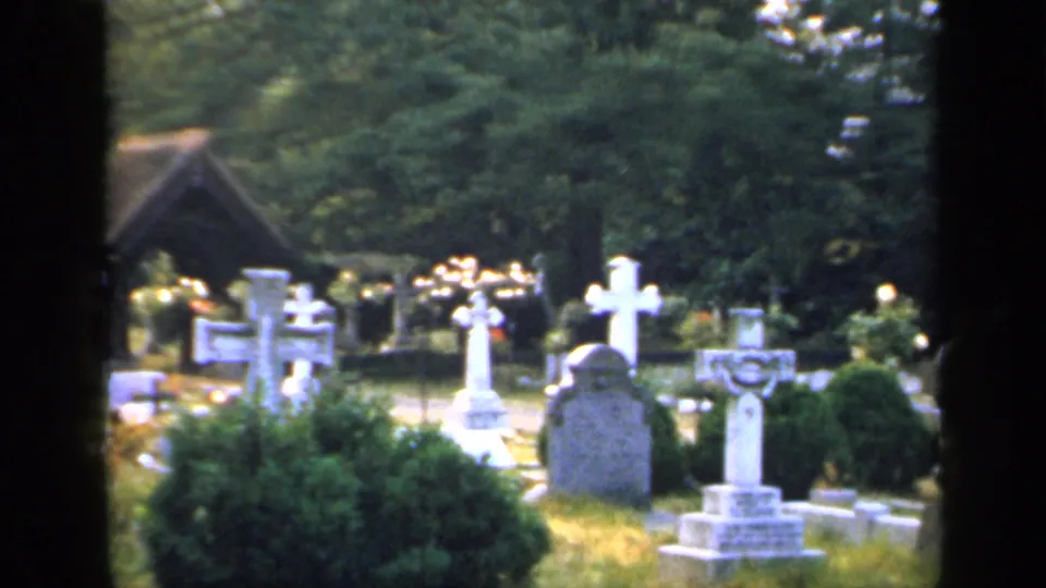 1949 England churchyard view with white cross markers, upright headstones, clipped shrubs, leafy trees, and a wooden shelter in the background.