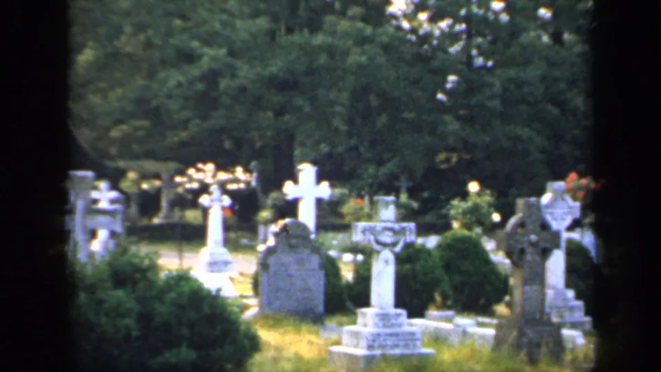 1949 England 8mm frame showing a cemetery with headstones, white crosses, shrubs, grass, and tree cover.
