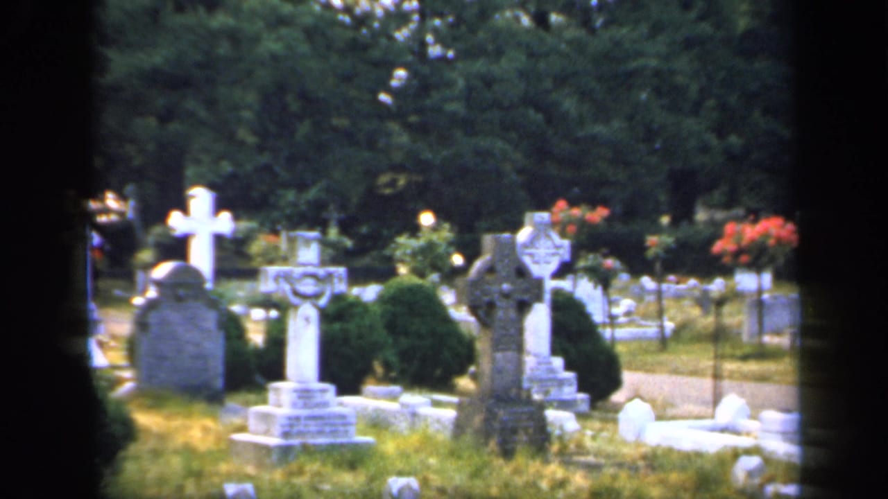 1949 England cemetery frame with stone crosses, rounded grave markers, clipped shrubs, grass, trees, and a visible path.