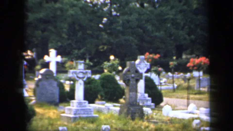 1949 England cemetery frame with stone crosses, rounded grave markers, clipped shrubs, grass, trees, and a visible path.
