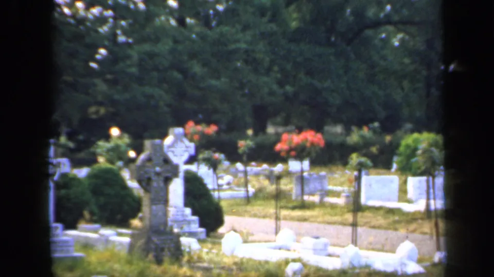 1949 England 8mm frame centered on a large stone cross with cemetery plots, flowers, shrubs, grass, and trees behind it.