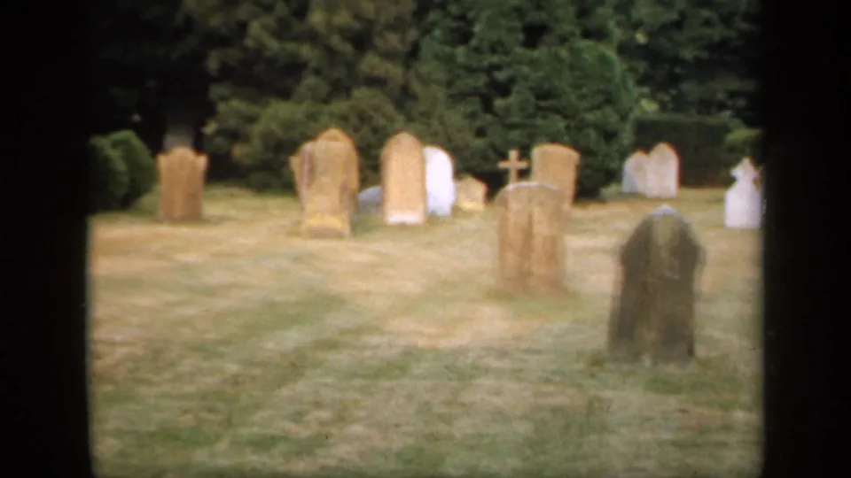 1949 England cemetery frame showing upright older headstones in dry grass, with dense trees and a small cross marker in the background.