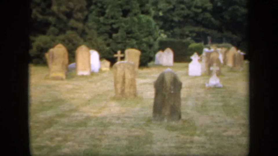 1949 England 8mm frame showing a quiet cemetery with aged upright gravestones, crosses, grass, hedges, and trees beyond.