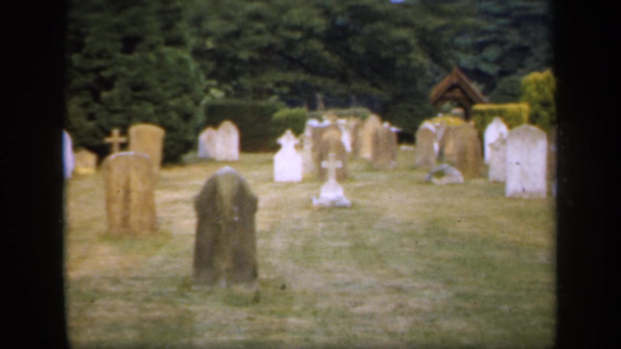 1949 England 8mm frame with a broad cemetery view, mixed stone headstones, white grave markers, cross monuments, and dense greenery.