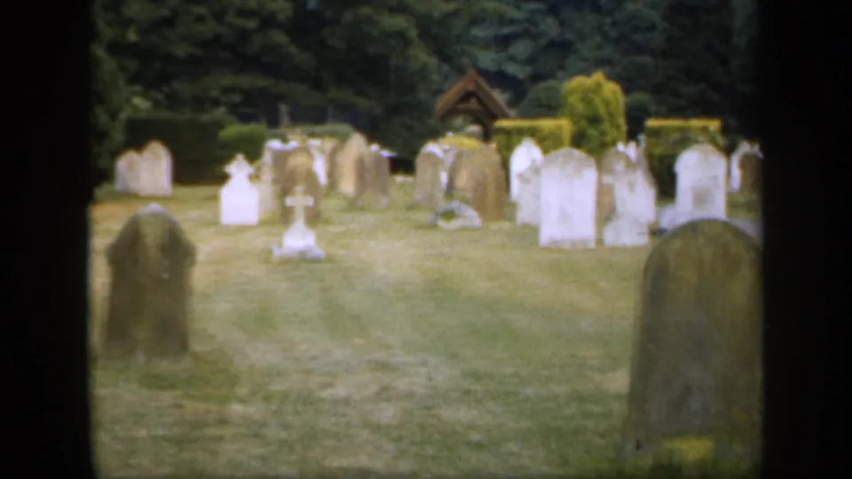 1949 England 8mm frame showing a soft, handheld view of a cemetery with foreground markers, pale headstones, trees, hedges, and a small timber structure.