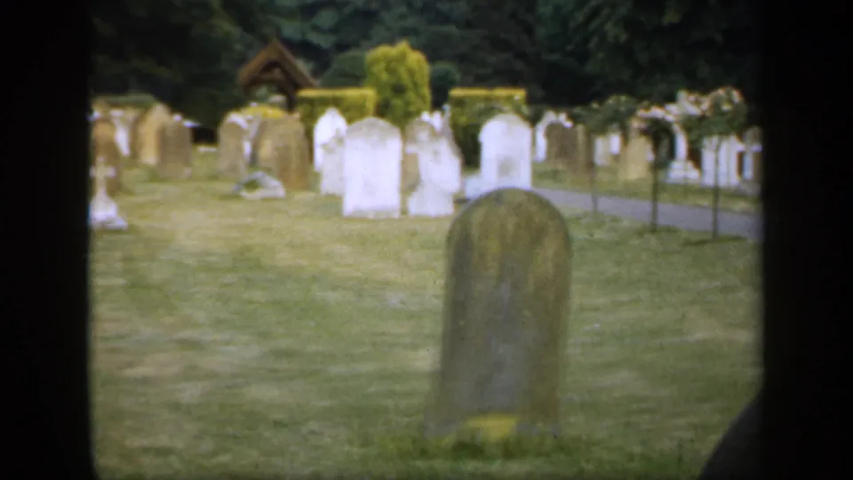 1949 England 8mm frame with a large rounded headstone in the foreground, scattered cemetery markers, crosses, hedges, and dark trees in the background.